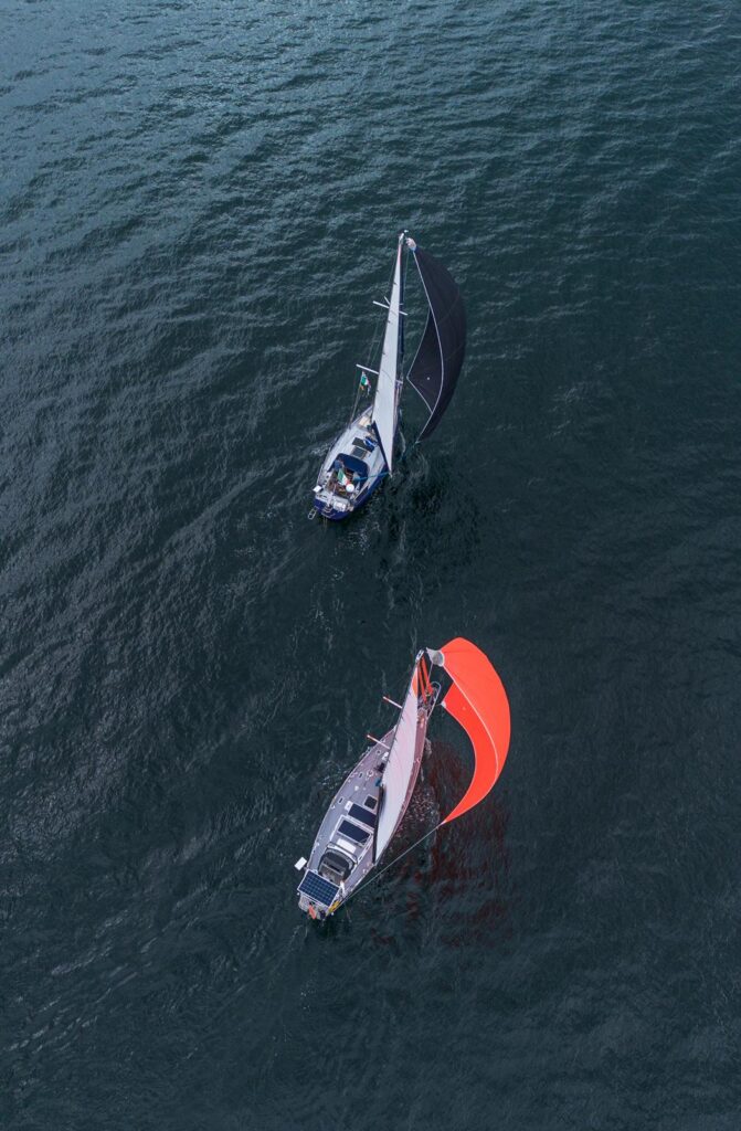 Aerial view of two yachts sailing downwind, one under a dark spinnaker and one under an orange spinnaker