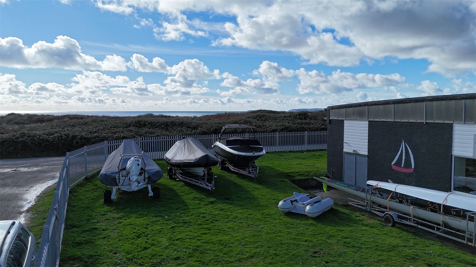 Covered boats on trailers in the secure storage compound beside the Boatshed Sailmakers loft in Pwllheli, with the sea behind