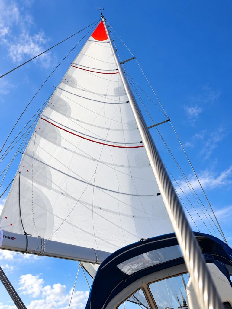 Looking up at a white cruising mainsail with full-length battens and a red head patch, above a navy sprayhood, against a blue sky