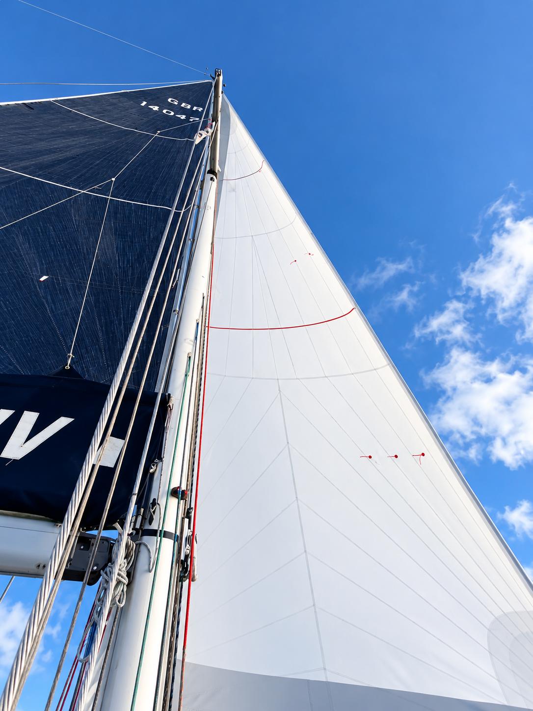 Looking up the mast at a dark mainsail with sail number GBR 14047 above its lazy bag, beside a white headsail against a blue sky